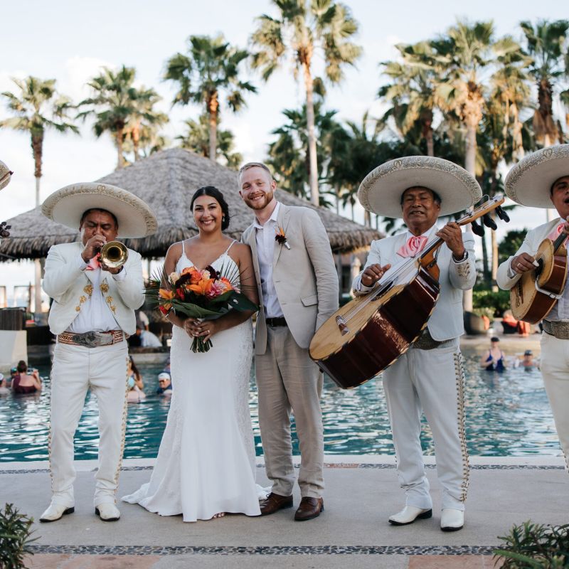 A bride and groom pose in a tropical scene with a mariachi band playing music around them. 